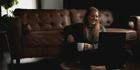 A female photographer sitting on the floor of a studio with a laptop and a mug, in front of a brown leather sofa.
