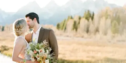 A bride and groom embracing in a field with a mountain range in the background.