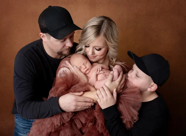 A family of four posing in a studio with a brown backdrop, holding a sleeping newborn.