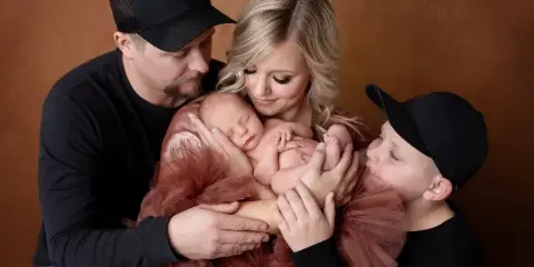 A family of four posing in a studio with a brown backdrop, holding a sleeping newborn.