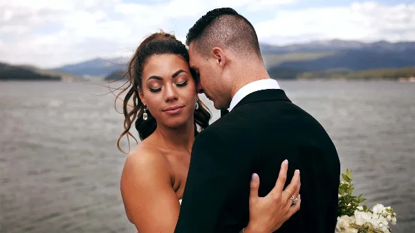 A cinematic close-up of a bride and groom embracing with their eyes closed, set against a blurred background of a lake and mountains.