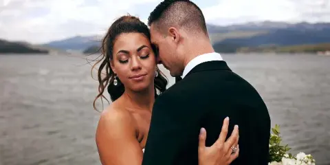 A cinematic close-up of a bride and groom embracing with their eyes closed, set against a blurred background of a lake and mountains.