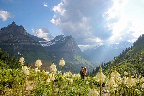 A bride and groom standing in a field of white wildflowers with a backdrop of rugged mountain peaks and a blue sky.