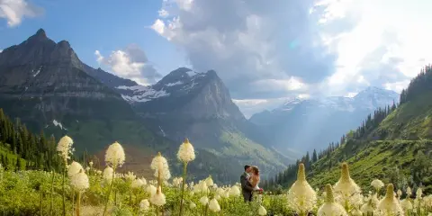 A bride and groom standing in a field of white wildflowers with a backdrop of rugged mountain peaks and a blue sky.