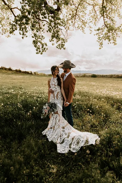 A bride in a white lace gown and a groom in a brown suit and cowboy hat standing in a grassy field with mountains in the background.