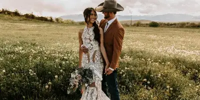 A bride in a white lace gown and a groom in a brown suit and cowboy hat standing in a grassy field with mountains in the background.