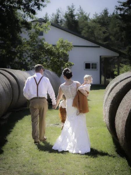 A bride and groom walking hand-in-hand with two young children in an outdoor setting near hay bales and a white barn.