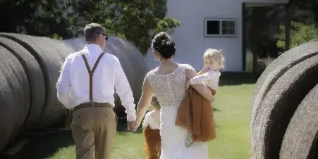 A bride and groom walking hand-in-hand with two young children in an outdoor setting near hay bales and a white barn.