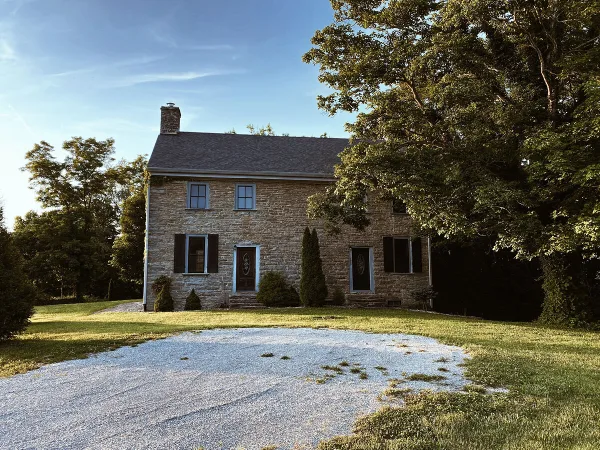 Exterior view of a historic two-story stone mansion with a gravel driveway and surrounding greenery.
