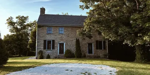 Exterior view of a historic two-story stone mansion with a gravel driveway and surrounding greenery.