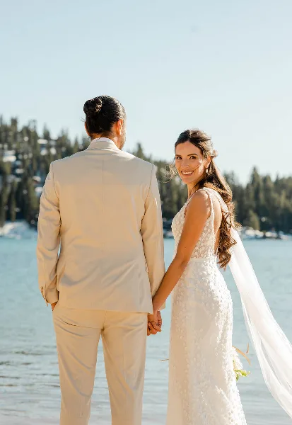 A bride and groom holding hands on a beach at Lake Tahoe, with the bride smiling at the camera and the groom looking out at the water.