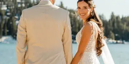 A bride and groom holding hands on a beach at Lake Tahoe, with the bride smiling at the camera and the groom looking out at the water.