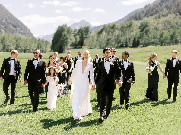 A wedding party consisting of the bride, groom, and attendants walking across a grassy field with mountains in the background.