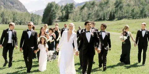 A wedding party consisting of the bride, groom, and attendants walking across a grassy field with mountains in the background.