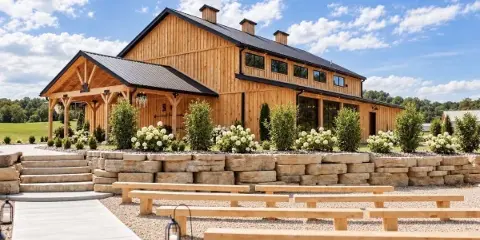 Exterior view of a large wooden wedding venue with a covered porch, stone retaining walls, and outdoor benches.