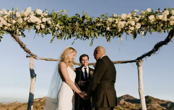 A bride and groom exchanging vows with an officiant under a floral arch during a wedding ceremony.