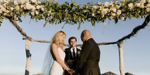 A bride and groom exchanging vows with an officiant under a floral arch during a wedding ceremony.