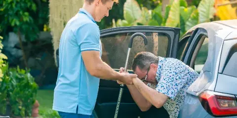 A driver in a blue polo shirt assisting a passenger with a cane into the back seat of a car.