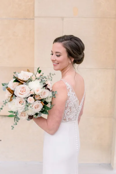 A bride in a white lace dress with an elegant updo hairstyle holding a bouquet of white and blush roses.