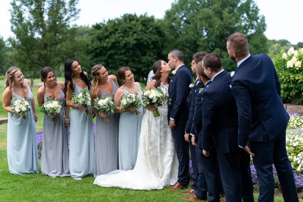 A wedding party consisting of a bride, groom, bridesmaids, and groomsmen standing outdoors on a lawn.