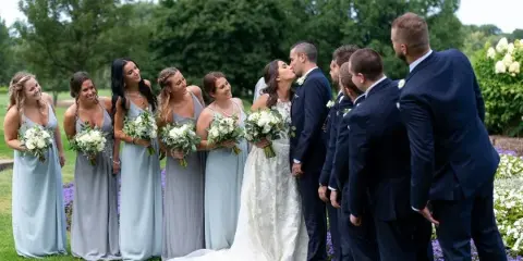A wedding party consisting of a bride, groom, bridesmaids, and groomsmen standing outdoors on a lawn.