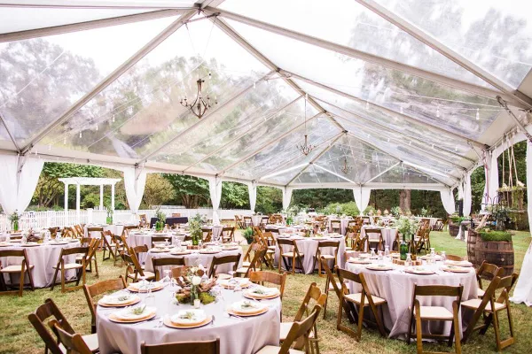 Interior view of a large clear-top wedding tent with round tables, wooden chairs, and hanging chandeliers.