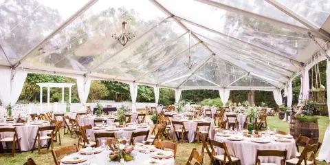 Interior view of a large clear-top wedding tent with round tables, wooden chairs, and hanging chandeliers.