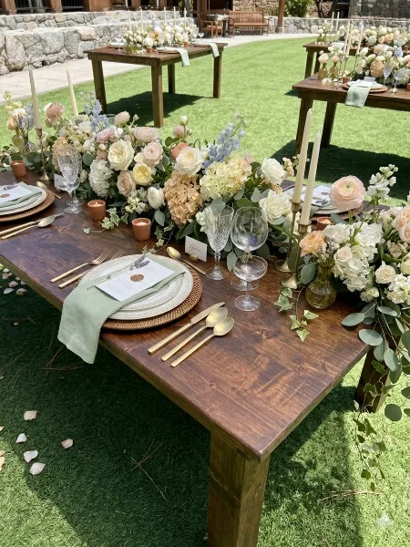 A rustic wooden farm table set with floral arrangements, candles, and gold cutlery on a lawn.