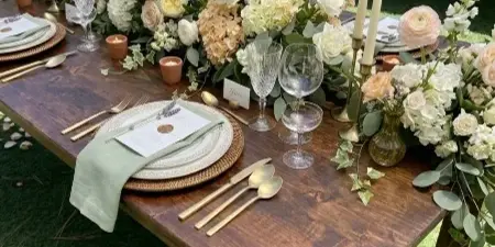 A rustic wooden farm table set with floral arrangements, candles, and gold cutlery on a lawn.