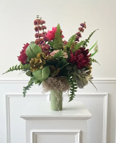 A tall floral arrangement with red and pink blooms, greenery, and ferns in a textured green vase on a white pedestal.