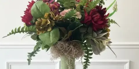 A tall floral arrangement with red and pink blooms, greenery, and ferns in a textured green vase on a white pedestal.