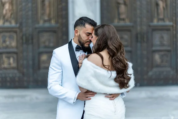 A professional wedding photograph showing a groom in a white tuxedo and a bride in a white dress and fur stole embracing in front of an ornate stone building.
