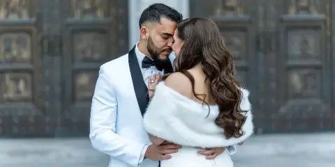A professional wedding photograph showing a groom in a white tuxedo and a bride in a white dress and fur stole embracing in front of an ornate stone building.