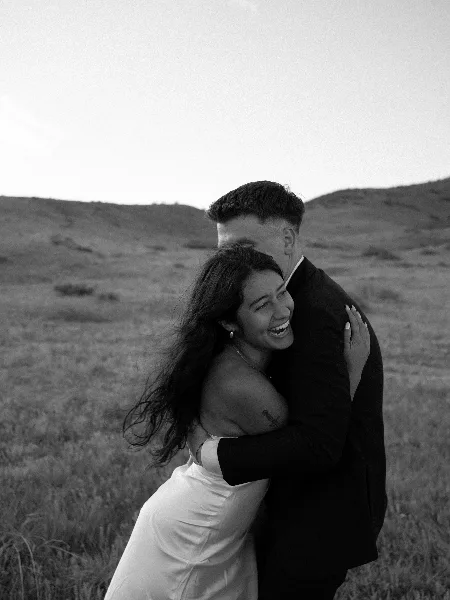 A black and white photograph of a bride and groom embracing in a grassy field.