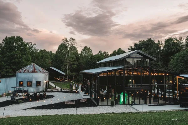 Wide exterior shot of a modern glass-walled greenhouse wedding venue and a rustic barn at dusk, featuring string lights and a gravel courtyard.