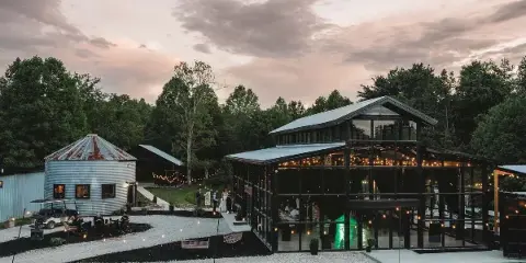 Wide exterior shot of a modern glass-walled greenhouse wedding venue and a rustic barn at dusk, featuring string lights and a gravel courtyard.