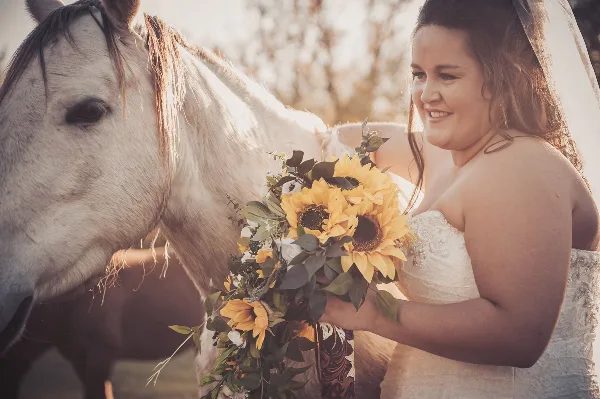 A bride in a white wedding dress and veil holding a sunflower bouquet while standing next to a white horse outdoors.