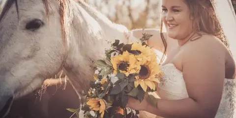 A bride in a white wedding dress and veil holding a sunflower bouquet while standing next to a white horse outdoors.