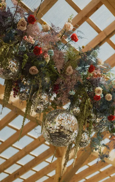 A vibrant, artistic floral installation with red and cream roses, wild greenery, and hanging disco balls suspended from a wooden pergola structure.