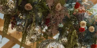 A vibrant, artistic floral installation with red and cream roses, wild greenery, and hanging disco balls suspended from a wooden pergola structure.
