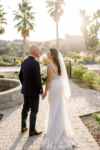A bride and groom holding hands and smiling at each other on a paved path with palm trees in the background.