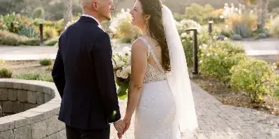 A bride and groom holding hands and smiling at each other on a paved path with palm trees in the background.
