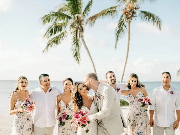 A wedding party posing on a tropical beach with palm trees and the ocean in the background.