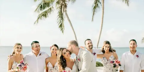 A wedding party posing on a tropical beach with palm trees and the ocean in the background.