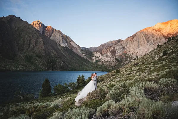 A bride and groom embracing on a hillside overlooking a mountain lake with rugged peaks in the background.