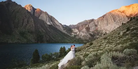 A bride and groom embracing on a hillside overlooking a mountain lake with rugged peaks in the background.
