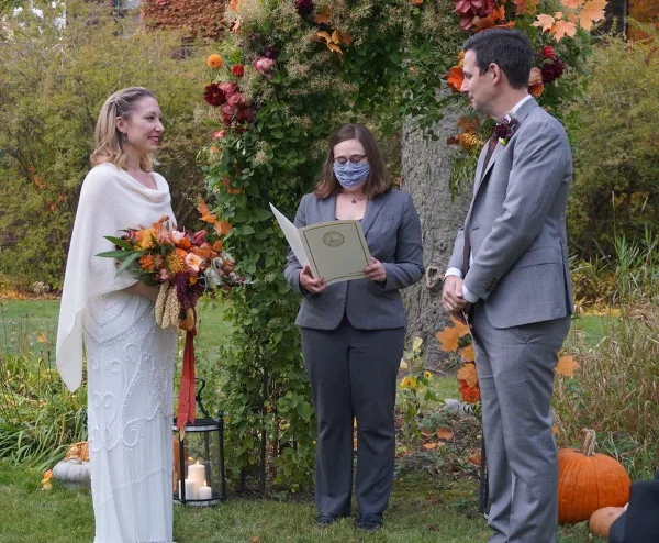 A wedding officiant wearing a face mask and holding a script stands between a bride and groom during an outdoor ceremony.