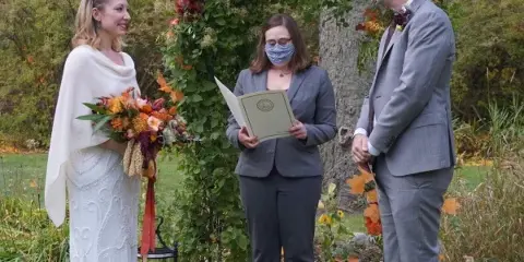 A wedding officiant wearing a face mask and holding a script stands between a bride and groom during an outdoor ceremony.
