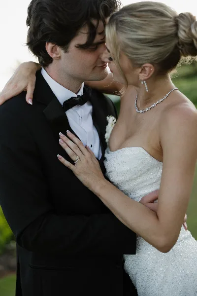 A bride with a styled updo and a groom in a tuxedo embracing outdoors.