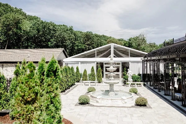 Outdoor wedding venue courtyard featuring a white tent, stone building, fountain, and manicured gardens.
